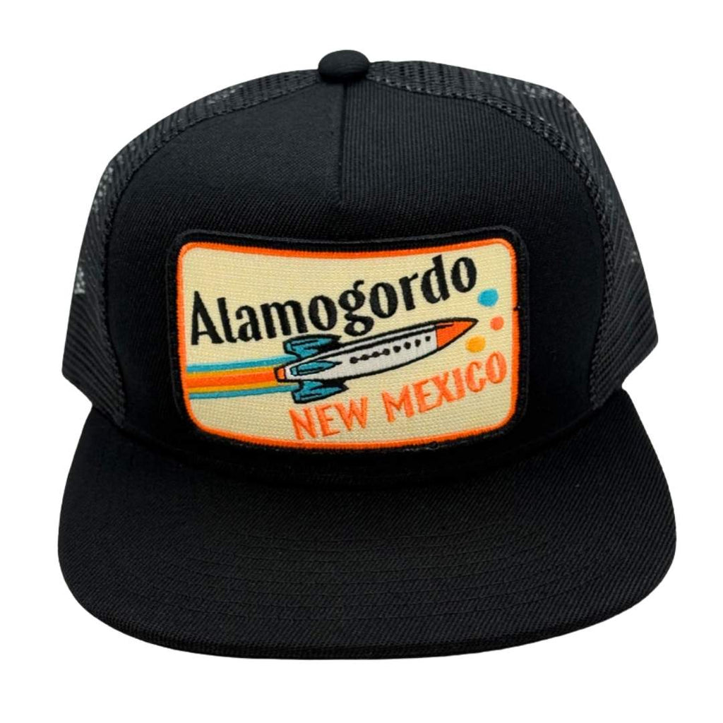 A black trucker hat with a rectangular embroidered patch on the front that reads 'Alamogordo New Mexico' with a retro airplane graphic, shown on a plain white background.
