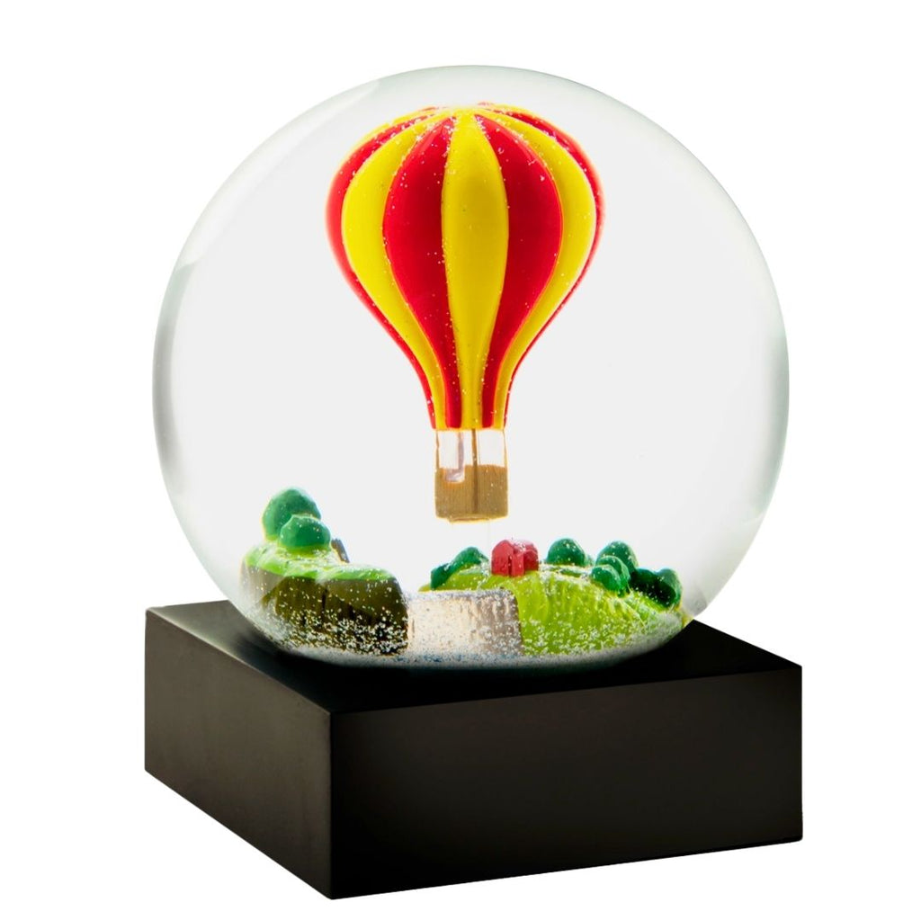 A clear glass snow globe on a black rectangular base, featuring a red and yellow striped hot air balloon floating above miniature green hills and trees.