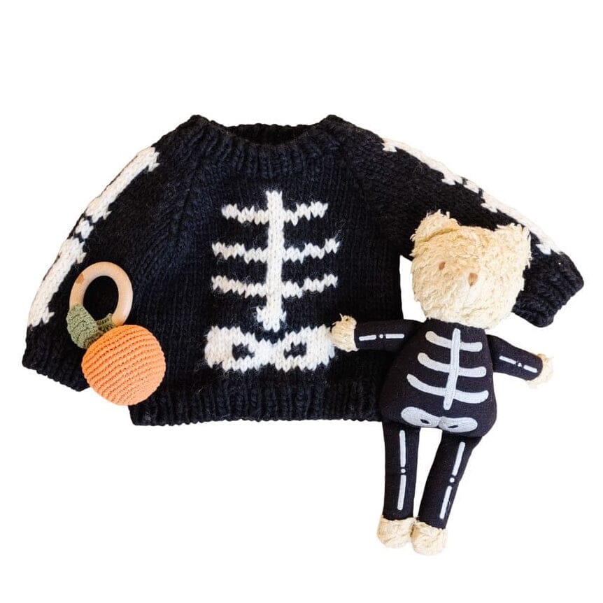 A black knit baby sweater with a white skeleton print, shown with a plush teddy bear doll wearing a matching black and white skeleton outfit and a small orange knit rattle on a white background.