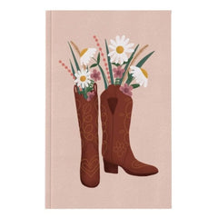 A notebook with a light pink cover notebook featuring an illustration of two brown cowboy boots filled with white daisies and wildflowers, shown against a plain white background.