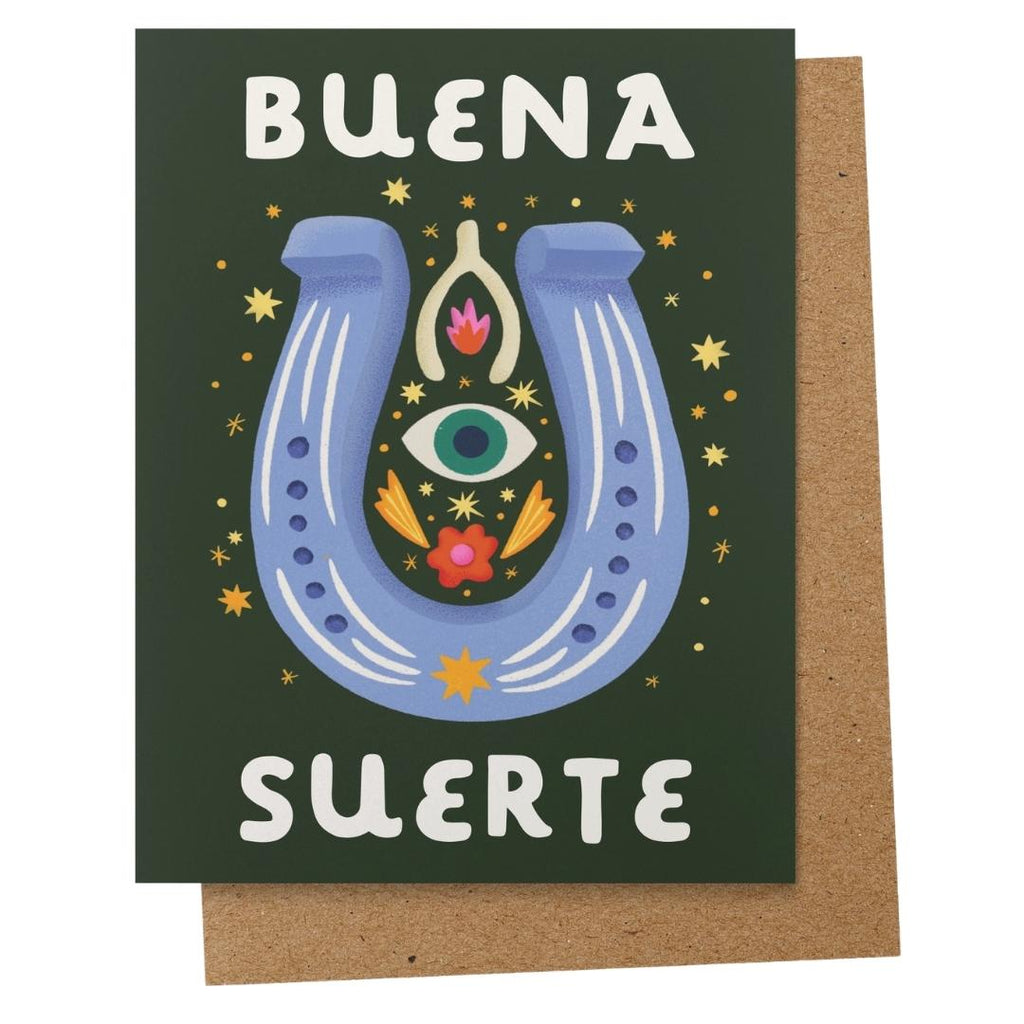 A dark green greeting card with a colorful illustration of a blue horseshoe, an all-seeing eye, and flowers, with the Spanish text 'BUENA SUERTE' in white. A brown kraft envelope is partially visible behind the card.