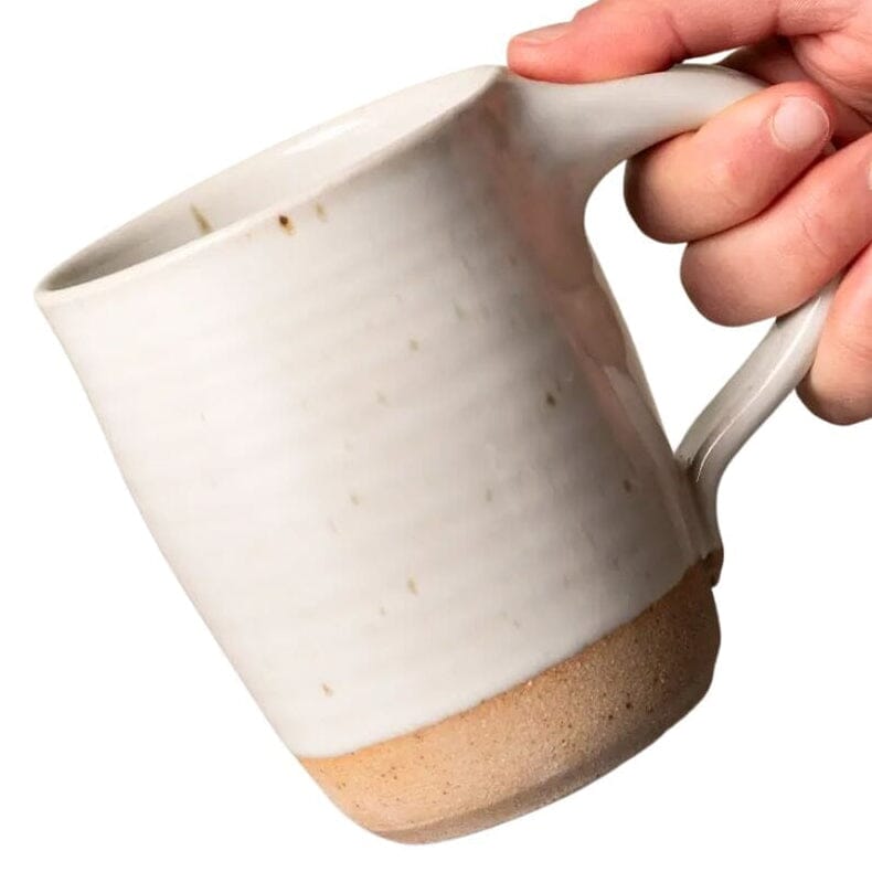 A hand holding a handmade ceramic mug with a textured off-white glazed body and a natural tan unglazed base, shown against a plain white background.