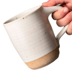 A hand holding a handmade ceramic mug with a textured off-white glazed body and a natural tan unglazed base, shown against a plain white background.