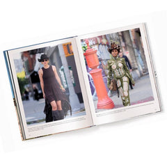 An open fashion book displayed on a white background, showing two full-page editorial photos. The left page features a model in a black dress and hat, and the right page shows a model in an ornate green and gold costume, with street scenes and small text below the images.