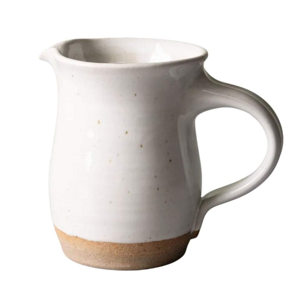 A small ceramic pitcher with a glossy off-white body, subtle speckles, and a natural tan unglazed base, shown on a plain white background.