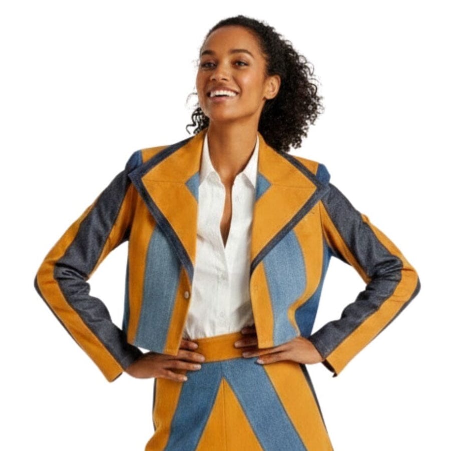 A woman modeling a colorful geometric-patterned blazer and matching skirt set. The outfit features bold stripes and angular panels of camel brown and denim styled with a white button-down shirt.