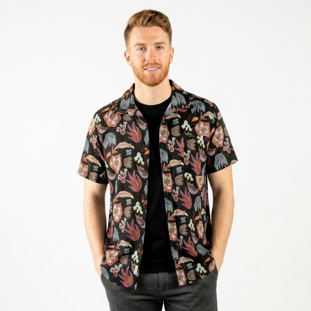 A man wearing a short-sleeve button-up shirt with a colorful Southwest patterns on a black background, layered over a black t-shirt and dark gray jeans, photographed against a solid white studio background.