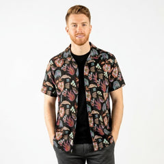 A man wearing a short-sleeve button-up shirt with a colorful Southwest patterns on a black background, layered over a black t-shirt and dark gray jeans, photographed against a solid white studio background.
