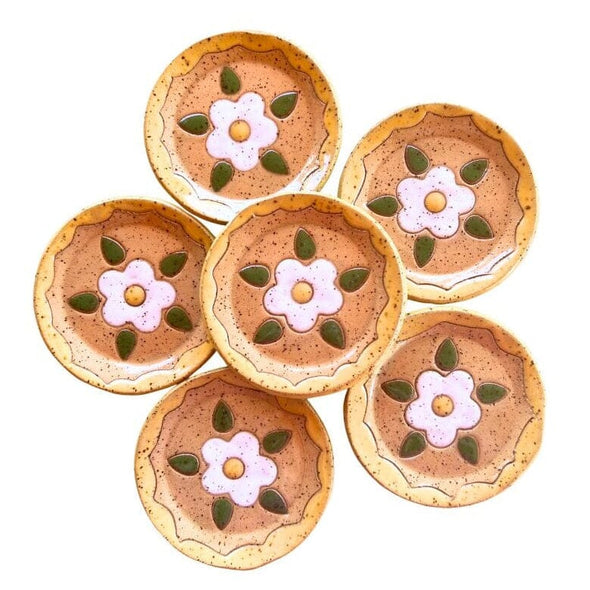Daisy round ceramic trinket dish arranged in a flower-like cluster on a white background. Each coaster has a speckled tan-brown base with a central pink flower motif, a yellow center, and green leaf accents.