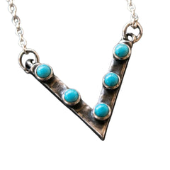 A silver-toned V-shaped pendant necklace featuring five round turquoise blue gemstones set along the arm of the letter V, shown against a plain white background.