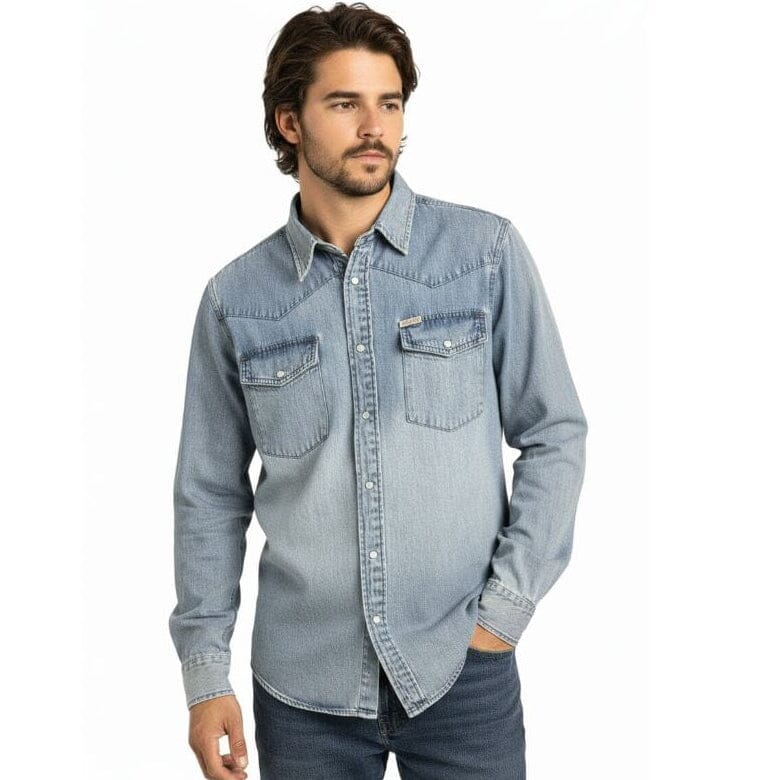 A man wearing a light blue denim western-style button-down shirt with two chest pockets, paired with dark blue jeans, against a plain white background.