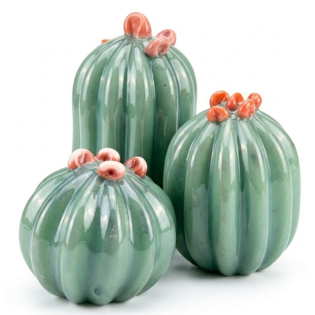 Three handblown glass ceramic cactus figurines in shades of sage green with small coral-pink tops, arranged on a white background.