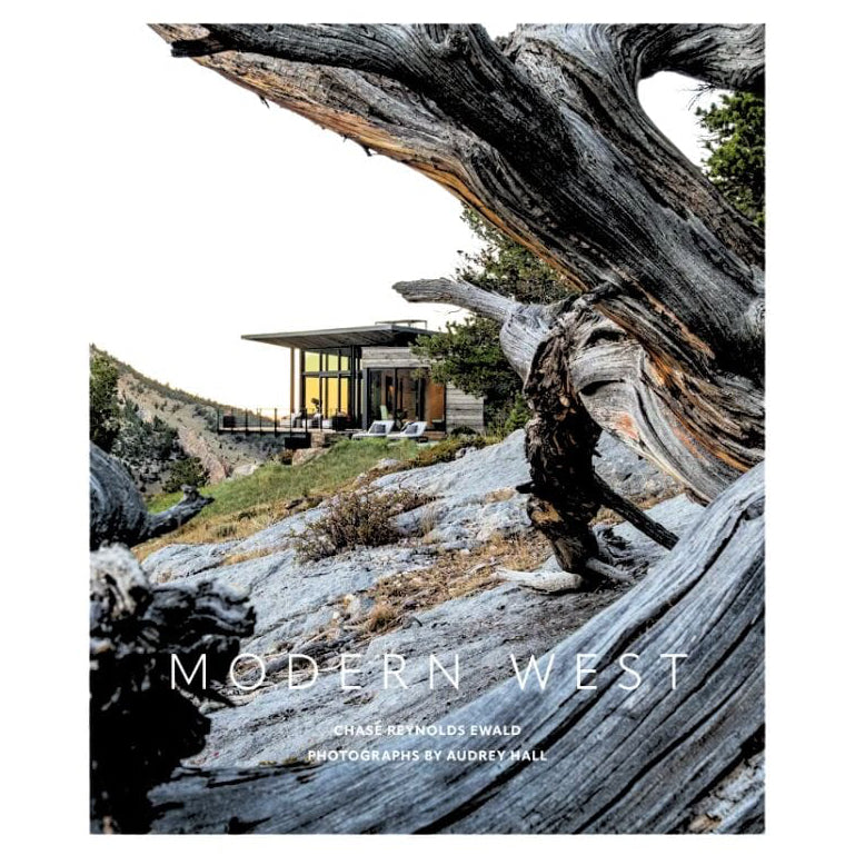 A book cover titled "MODERN WEST" with a photograph of a modern house on a rocky hillside, framed by large weathered tree trunks in the foreground. The cover has a white border and visible text reading "CHASE REYNOLDS Ewald" and "Photographs by Audrey Hall.