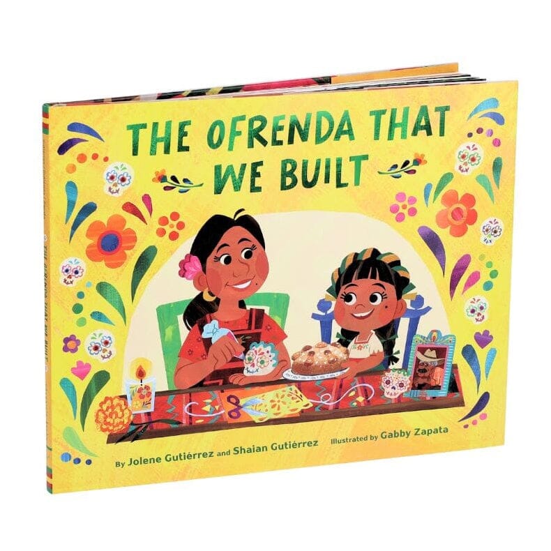 The Ofrenda That We Built children's book shown at a slight angle on a plain white background. The cover is bright yellow with colorful illustrated flowers, two children at a table, and festive decorative motifs.