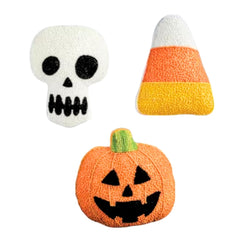 Three Halloween-themed decorative items on a plain white background: a white skull with black embroidered details, a candy corn piece with white, orange, and yellow sections, and an orange jack-o'-lantern with black facial features and a small green stem.