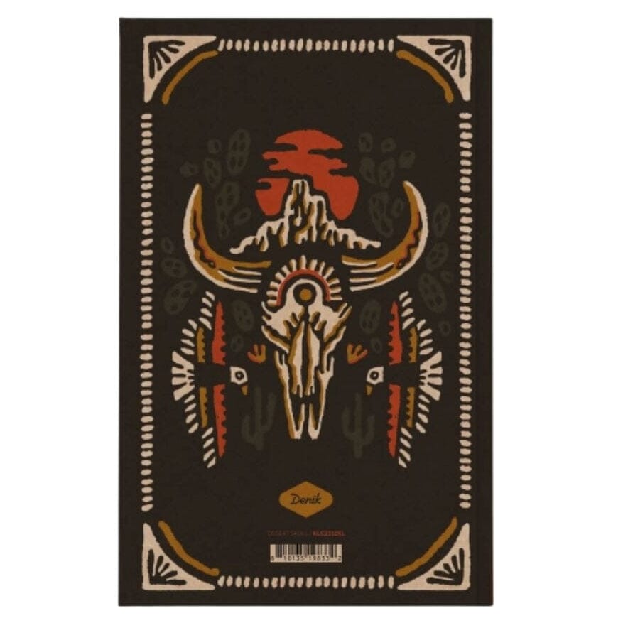 A rectangular dark brown rug with a central Native American-inspired design featuring a skull, curved horns, cacti, geometric borders, and an orange-red sun motif. The brand name 'Dusk' is printed near the bottom center.