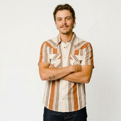A man with a mustache and short brown hair stands with arms crossed, wearing a short-sleeve button-up shirt with vertical stripes in orange, beige, and brown, paired with dark blue jeans, against a plain white background.