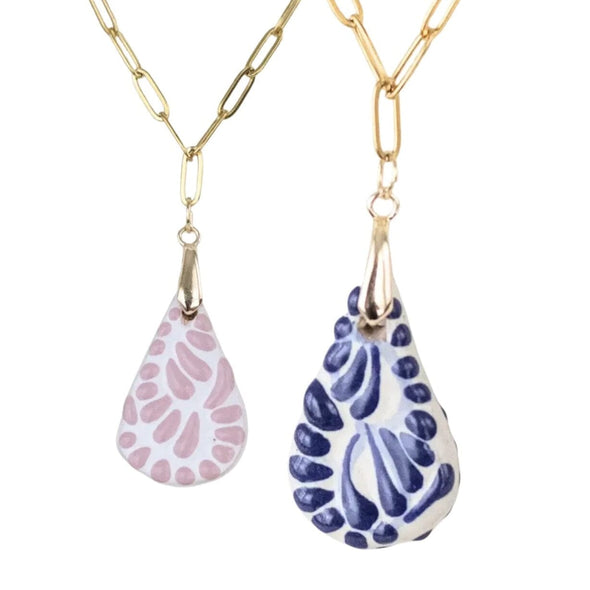 Two Talavera ceramic teardrop-shaped pendant necklaces on gold paperclip chains against a white background. One pendant is white with a pink abstract pattern, and the other is white with a dark blue abstract pattern.