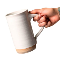 A hand holding a handmade ceramic tall mug with a textured off-white glazed body and a natural tan unglazed base, shown against a plain white background.
