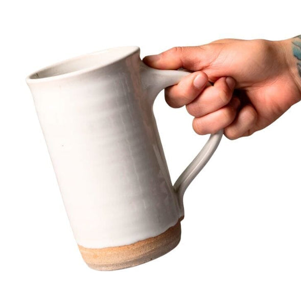 A hand holding a handmade ceramic tall mug with a textured off-white glazed body and a natural tan unglazed base, shown against a plain white background.