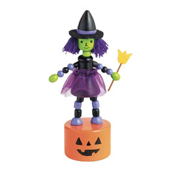 A small Halloween witch toy figurine standing on an orange jack-o'-lantern base. The figure has a green face and hair, a black hat, purple wig, black dress, purple tutu, green gloves, and holds a yellow broom.
