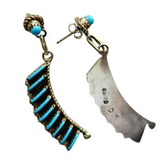 A pair of turquoise dangle earrings by Davis Kaamasee on a plain white background. One earring features a curved gold-toned setting with multiple bright turquoise-blue stones, and the other features a scalloped silver-toned metal charm with his hallmark.
