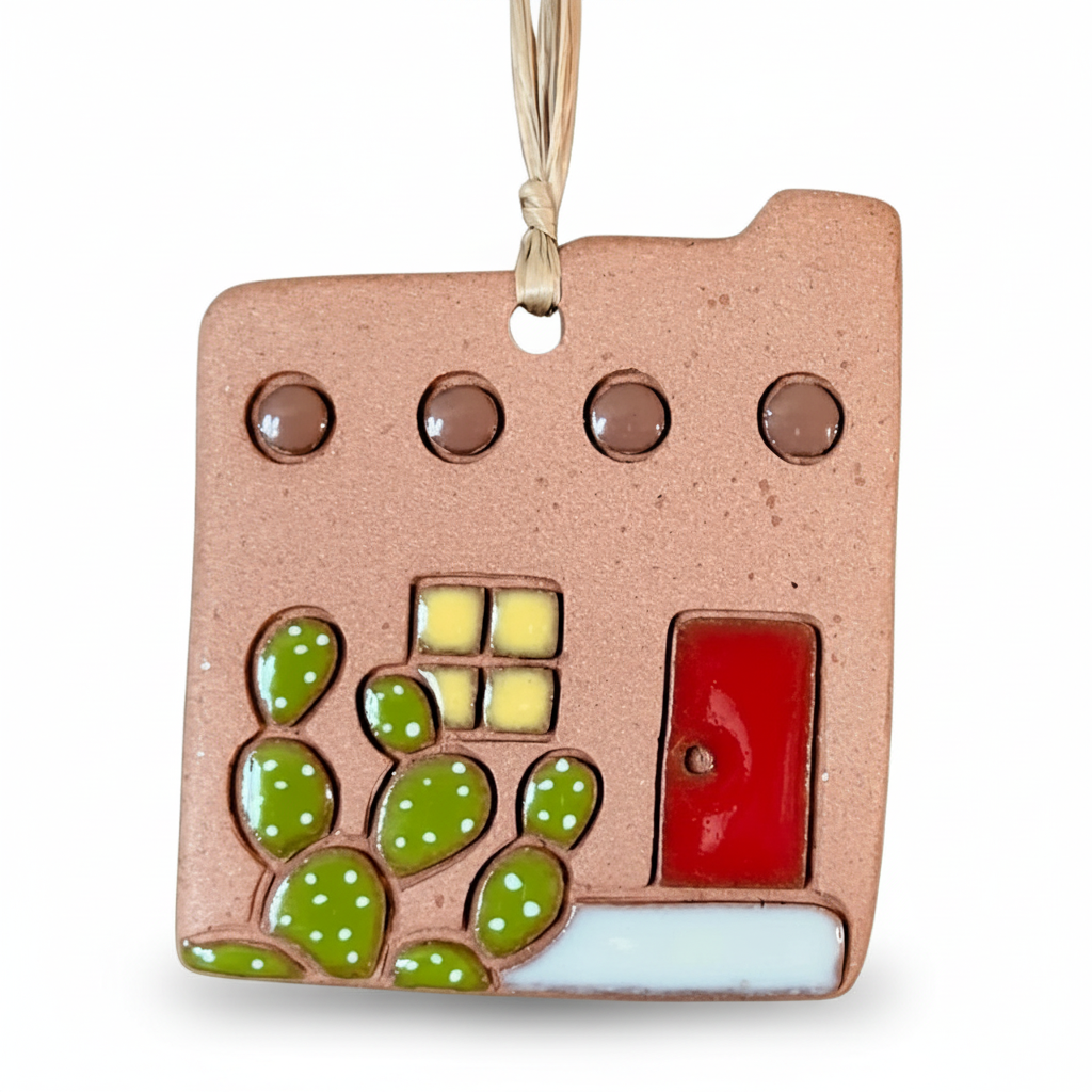 A square ceramic ornament shaped like an adobe house with a hanging loop at the top. The house has a warm terracotta base, a red door and a green cactus all on a clean white background.