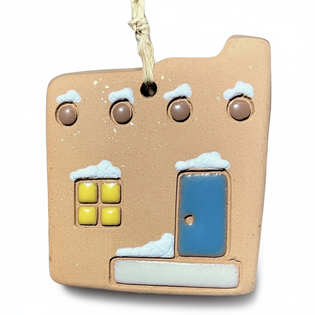 A small adobe house-shaped ornament in a warm beige tone, with a blue door, white snow details, and yellow window panes, hanging by a twisted string on a white background.
