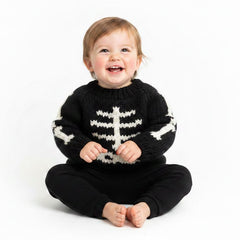 A smiling toddler sitting cross-legged on a white studio background, wearing a black knit sweater with a white skeleton motif and black pants.