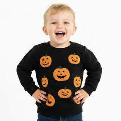 A young toddler boy wearing a black long-sleeve crewneck sweatshirt with a pattern of orange jack-o'-lantern pumpkins, paired with blue jeans and black shoes, standing against a solid white background.
